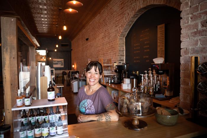 Artisan Mark owner Elayne Woods Jones stands behind the coffeeshop's counter. 在她身后, 很长一段, exposed brick wall extends to seating at the back of the shop, with warm lights overhead.